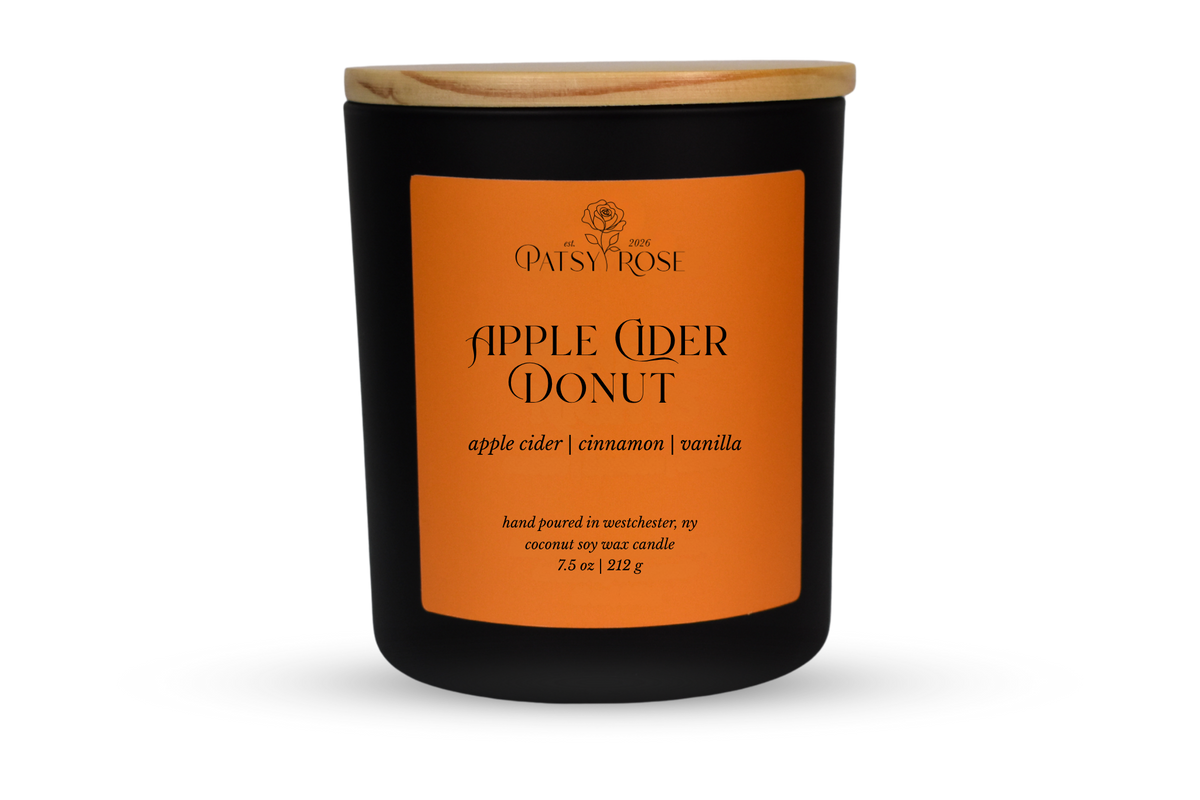 A 7.5 oz soy wax candle by Patsy Rose in the 'Apple Cider Donut' 'apple cider | cinnamon | vanilla' scent. The candle is in a matte black glass jar with a wood lid and an orange label, set against a plain white background.