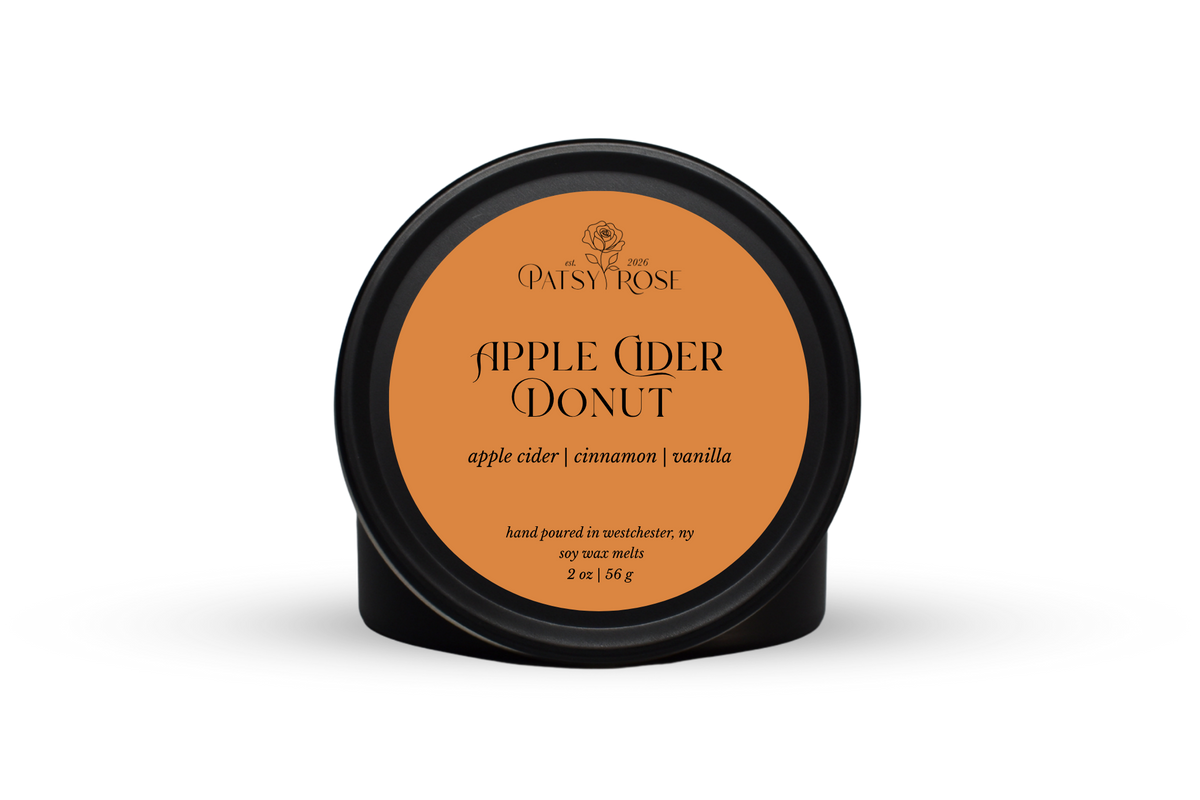 Hand-poured soy wax melts made by Patsy Rose with black tins and an orange label that says "Apple Cider Donut" with scent notes of "apple cider, cinnamon, vanilla".