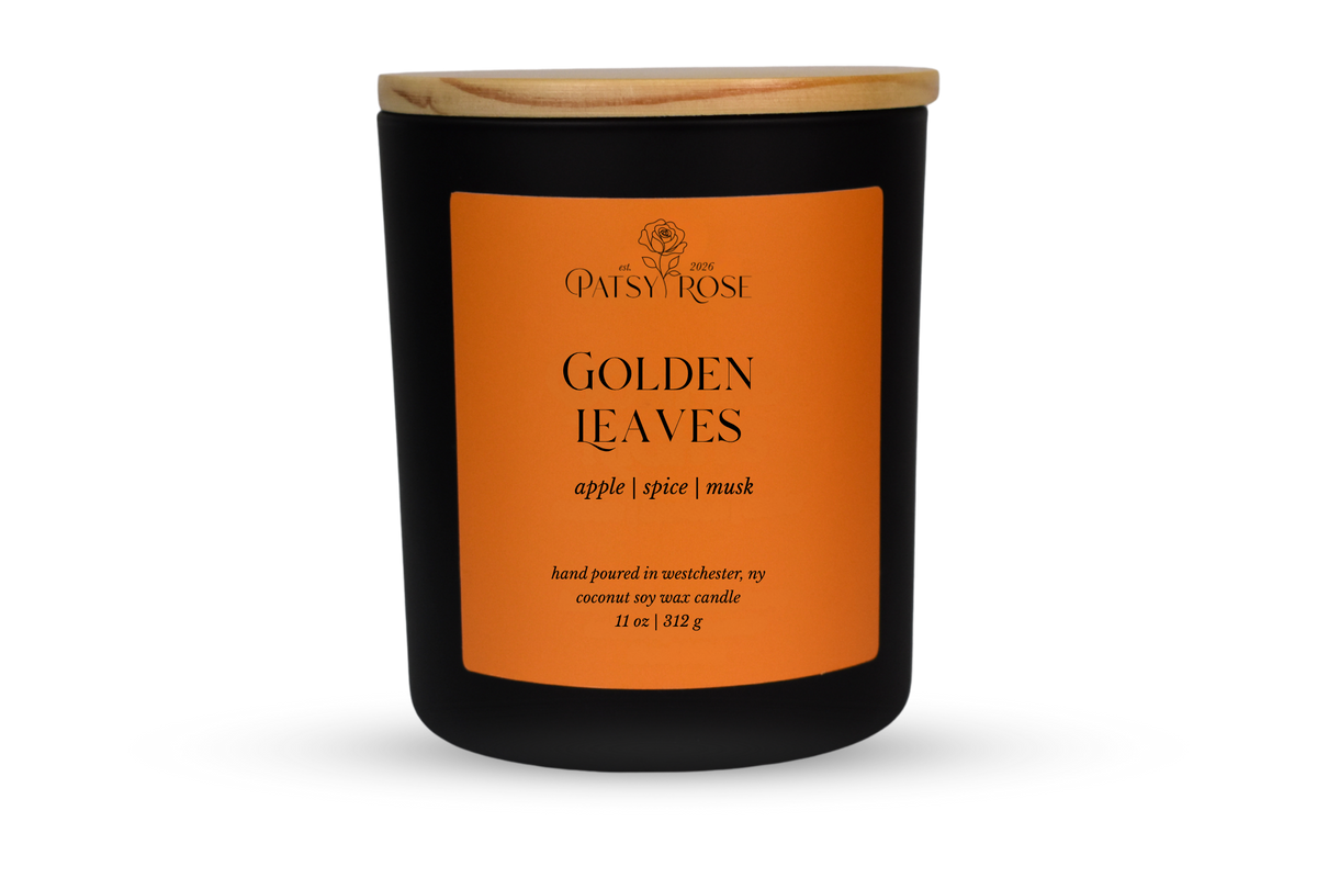 A 11 oz soy wax candle by Patsy Rose in the 'Golden Leaves' 'apple | spice | musk' scent. The candle is in a matte black glass jar with a wood lid and a orange label, set against a plain white background.
