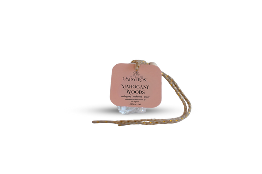 A peach-colored paper hang tag with black text reading 'Patsy Rose Mahogany Woods' on a glass rose shaped car air freshener, with a braided string attached, isolated on a white background.