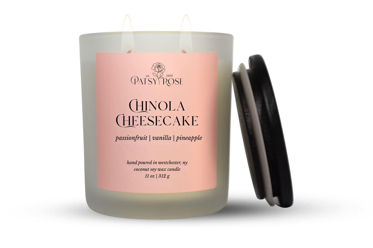 An 11 oz double-wick candle in a frosted glass jar with a pale pink label reading 'Patsy Rose Chinola Cheesecake' and 'passionfruit | vanilla | pineapple.' A black lid is leaning against the right side of the jar on a clean white background.