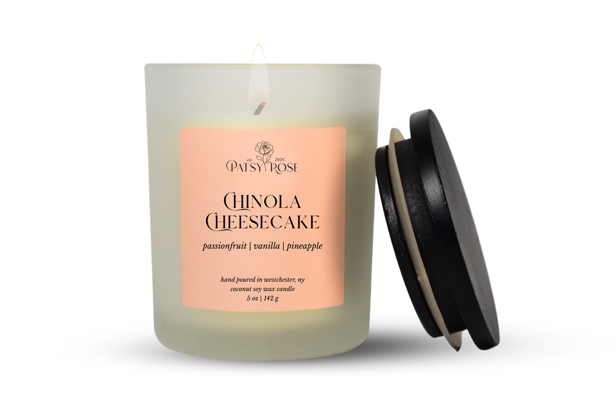 A single-wick coconut soy candle in a frosted glass jar with a pale pink label reading 'Patsy Rose Chinola Cheesecake' and 'passionfruit | vanilla | pineapple.' A black lid leans against the right side of the jar on a clean white background.