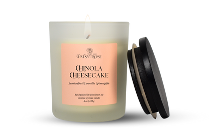 A single-wick coconut soy candle in a frosted glass jar with a pale pink label reading 'Patsy Rose Chinola Cheesecake' and 'passionfruit | vanilla | pineapple.' A black lid leans against the right side of the jar on a clean white background.