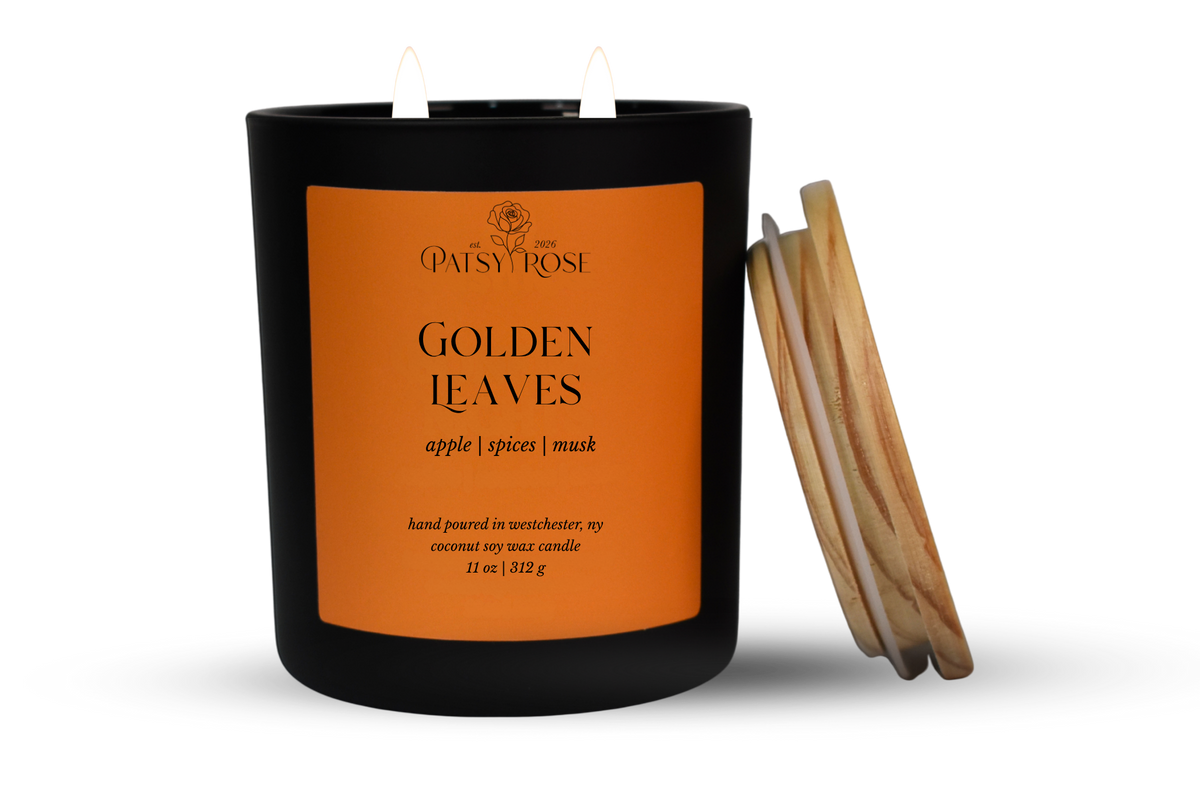 An 11 oz double-wick candle in a matte black glass jar with an orange label reading 'Patsy Rose Golden Leaves' and 'apple | spices | musk.' A wood lid is leaning against the right side of the jar on a clean white background.