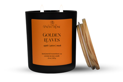 An 11 oz double-wick candle in a matte black glass jar with an orange label reading 'Patsy Rose Golden Leaves' and 'apple | spices | musk.' A wood lid is leaning against the right side of the jar on a clean white background.