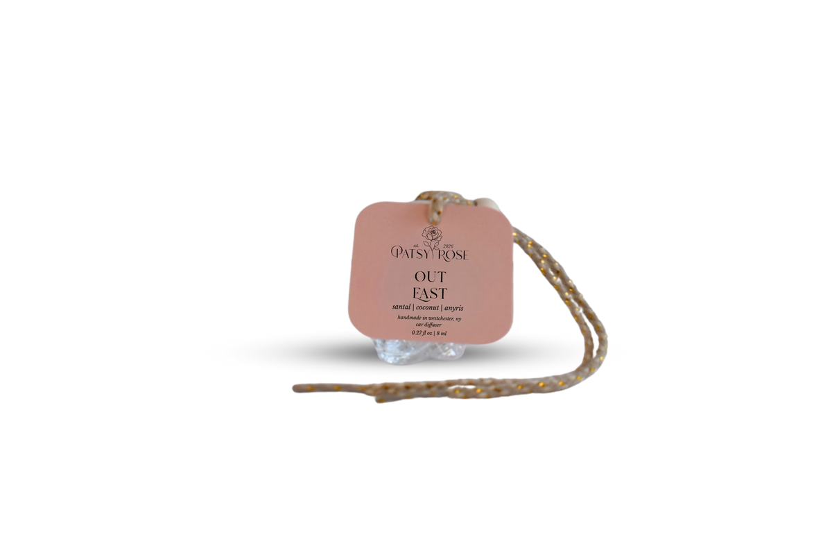 A peach-colored paper hang tag with black text reading 'Patsy Rose Out East' on a glass rose shaped car air freshener, with a braided string attached, isolated on a white background.