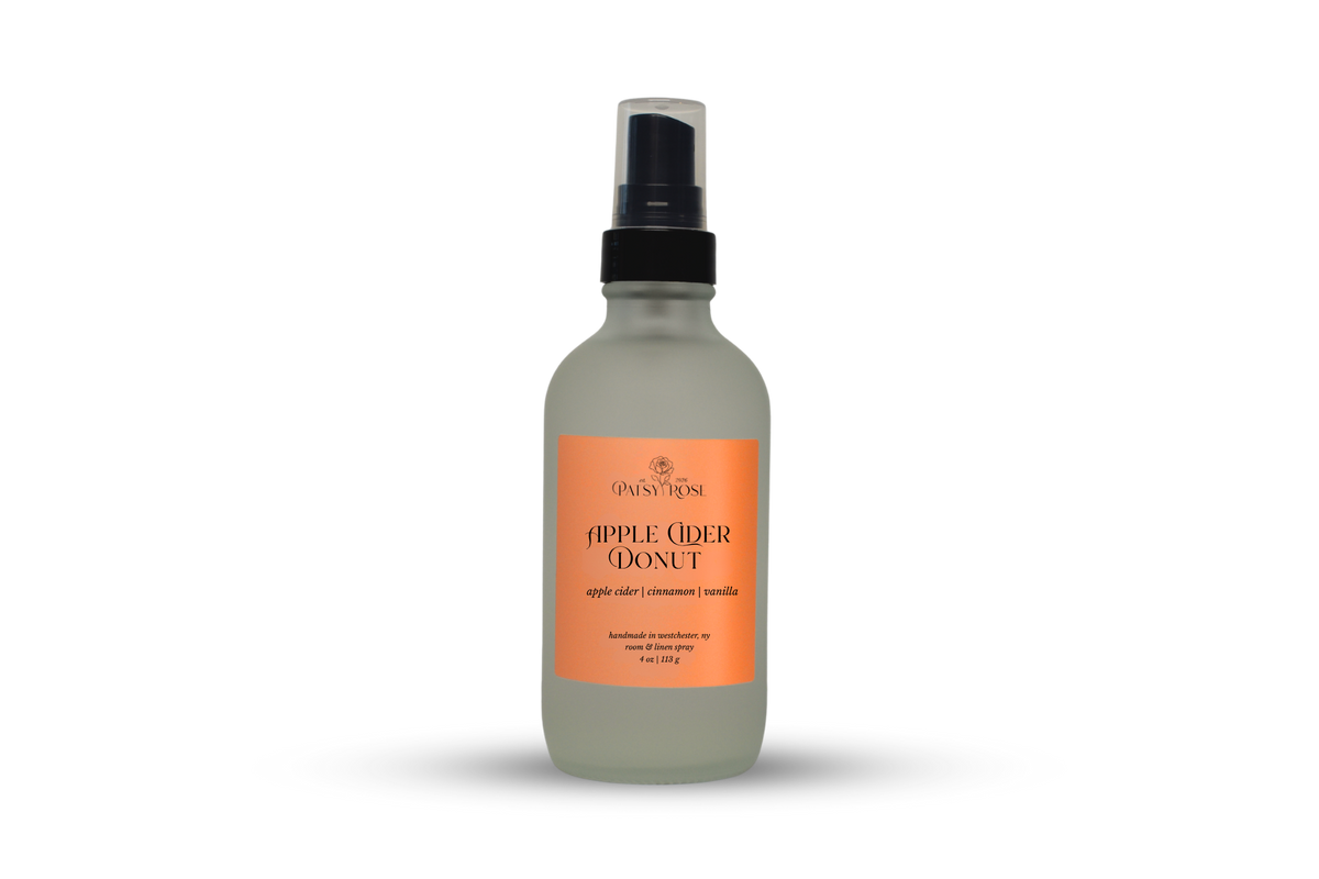 A 4 oz room and linen spray made by Patsy Rose in a frosted glass bottle with a peach label that says "Apple Cider Donut" with a scent of "apple cider, cinnamon, vanilla" on a white background