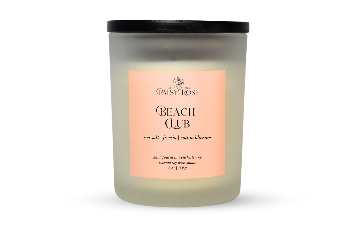 A 5 oz soy wax candle by Patsy Rose in the 'Beach Club' 'sea salt | freesia| cotton blossom' scent. The candle is in a frosted glass jar with a black lid and a peach-pink label, set against a plain white background.
