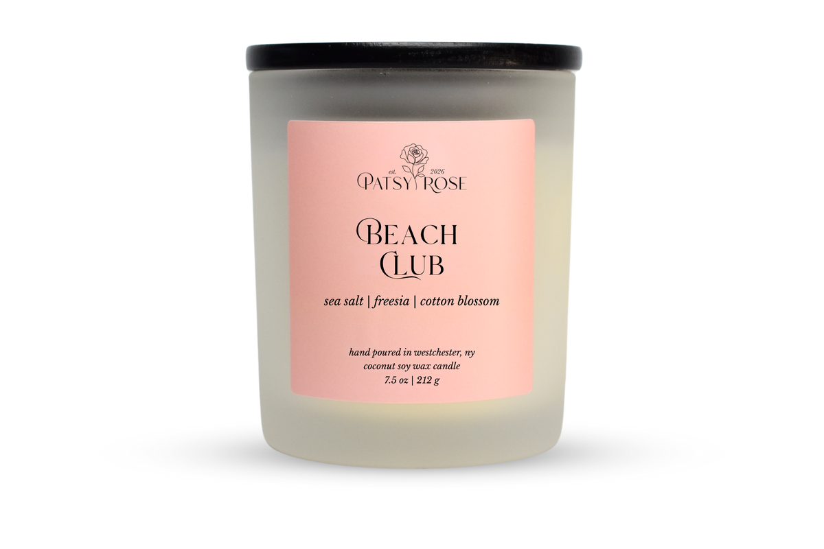 A 7.5 oz soy wax candle by Patsy Rose in the 'Beach Club' 'sea salt | freesia| cotton blossom' scent. The candle is in a frosted glass jar with a black lid and a peach-pink label, set against a plain white background.