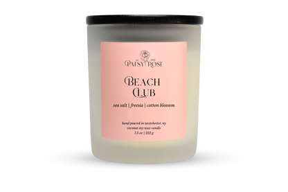 A 7.5 oz soy wax candle by Patsy Rose in the 'Beach Club' 'sea salt | freesia| cotton blossom' scent. The candle is in a frosted glass jar with a black lid and a peach-pink label, set against a plain white background.
