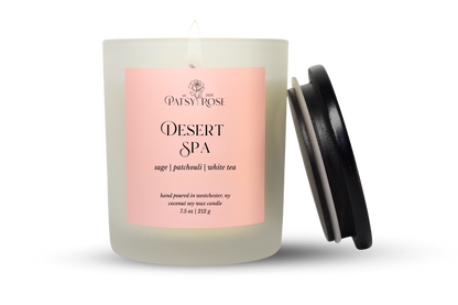 A 7.5 oz single-wick candle in a frosted glass jar with a pale pink label reading 'Patsy Rose Desert Spa' and 'sage | patchouli | white tea.' A black lid is leaning against the right side of the jar on a clean white background.