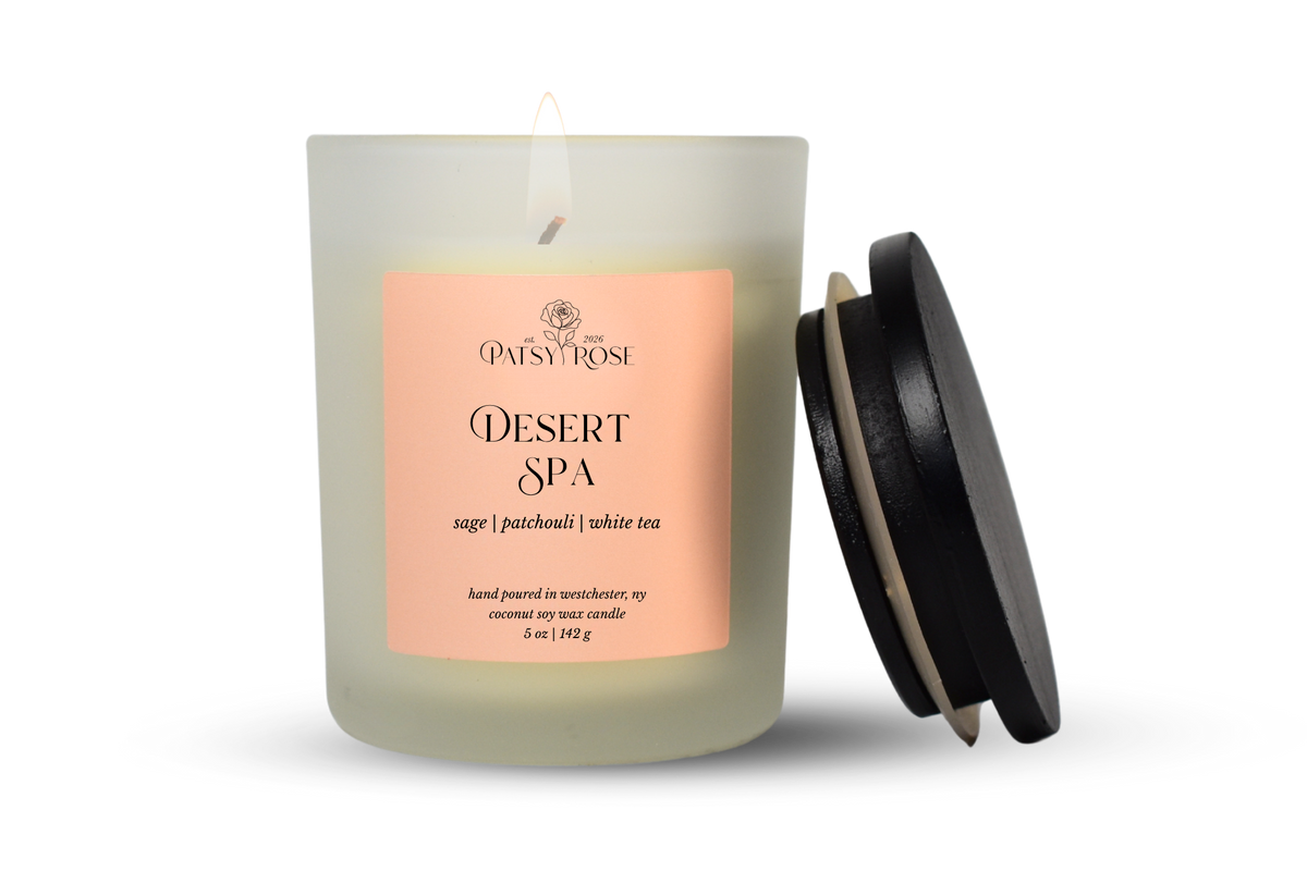 A single-wick coconut soy candle in a frosted glass jar with a pale pink label reading 'Patsy Rose Desert Spa' and 'sage | patchouli | white tea.' A black lid leans against the right side of the jar on a clean white background.