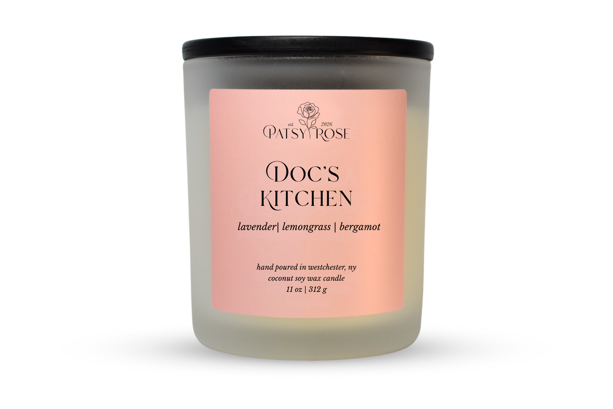 A 11 oz soy wax candle by Patsy Rose in the 'Doc's Kitchen' 'lavender | lemongrass | bergamot' scent. The candle is in a frosted glass jar with a black lid and a peach-pink label, set against a plain white background.