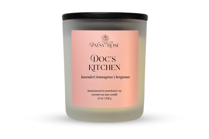 A 11 oz soy wax candle by Patsy Rose in the 'Doc's Kitchen' 'lavender | lemongrass | bergamot' scent. The candle is in a frosted glass jar with a black lid and a peach-pink label, set against a plain white background.
