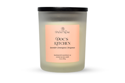 A 5 oz soy wax candle by Patsy Rose in the 'Doc's Kitchen' 'lavender| lemongrass | bergamot' scent. The candle is in a frosted glass jar with a black lid and a peach-pink label, set against a plain white background.