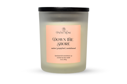 A 5 oz soy wax candle by Patsy Rose in the 'Down The Shore' 'melon| grapefruit | sandalwood' scent. The candle is in a frosted glass jar with a black lid and a peach-pink label, set against a plain white background.
