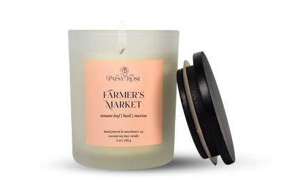A single-wick coconut soy candle in a frosted glass jar with a pale pink label reading 'Patsy Rose Farmer's Market' and 'tomato leaf | basil | marine.' A black lid leans against the right side of the jar on a clean white background.