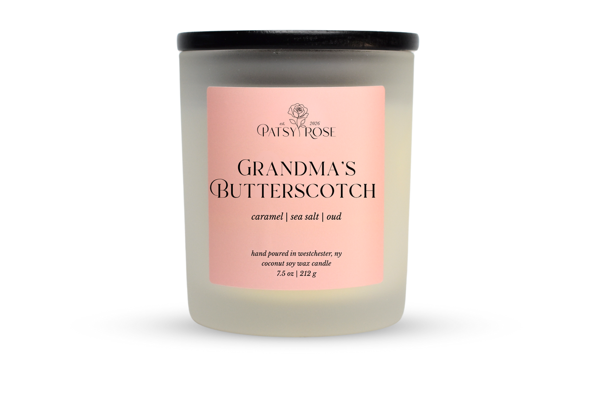 A 7.5 oz soy wax candle by Patsy Rose in the 'Grandma's Butterscotch' 'caramel | sea salt | oud' scent. The candle is in a frosted glass jar with a black lid and a peach-pink label, set against a plain white background.