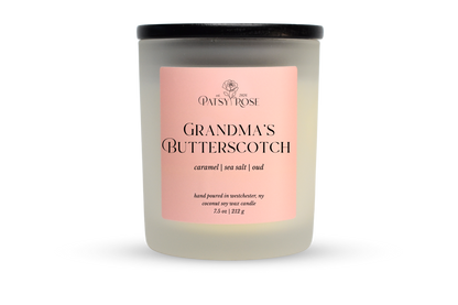A 7.5 oz soy wax candle by Patsy Rose in the 'Grandma's Butterscotch' 'caramel | sea salt | oud' scent. The candle is in a frosted glass jar with a black lid and a peach-pink label, set against a plain white background.