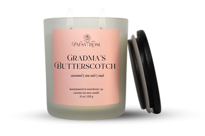 An 11 oz double-wick candle in a frosted glass jar with a pale pink label reading 'Patsy Rose Grandma's Butterscotch' and 'caramel | sea salt | oud.' A black lid is leaning against the right side of the jar on a clean white background.