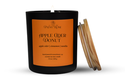 A single-wick coconut soy candle in a matte black glass jar with an orange label reading 'Patsy Rose Apple Cider Donut' and 'apple cider | cinnamon | vanilla.' A wood lid leans against the right side of the jar on a clean white background.