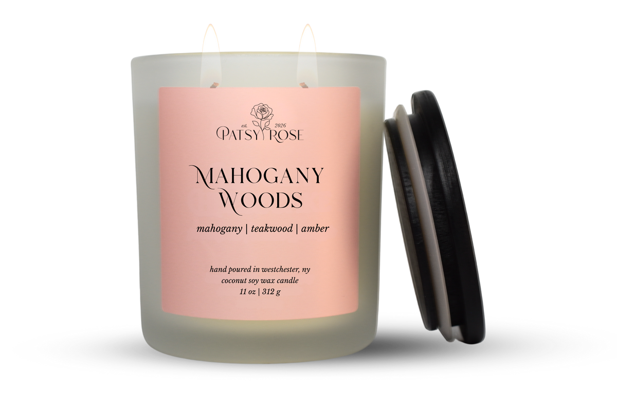 An 11 oz double-wick candle in a frosted glass jar with a pale pink label reading 'Patsy Rose Mahogany Woods' and 'mahogany | teakwood | amber.' A black lid is leaning against the right side of the jar on a clean white background.