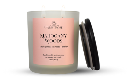 An 11 oz double-wick candle in a frosted glass jar with a pale pink label reading 'Patsy Rose Mahogany Woods' and 'mahogany | teakwood | amber.' A black lid is leaning against the right side of the jar on a clean white background.