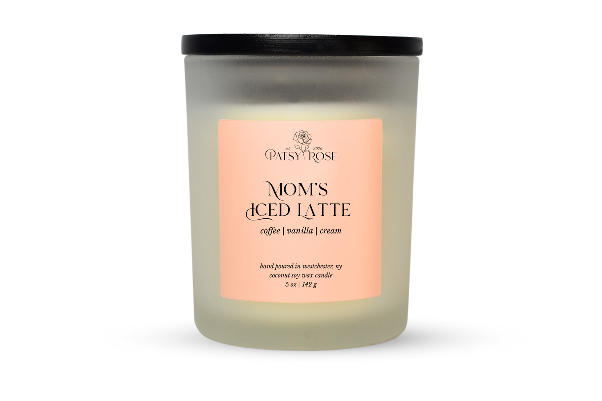 A 5 oz soy wax candle by Patsy Rose in the 'Mom's Iced Latte' 'coffee | vanilla | cream' scent. The candle is in a frosted glass jar with a black lid and a peach-pink label, set against a plain white background.