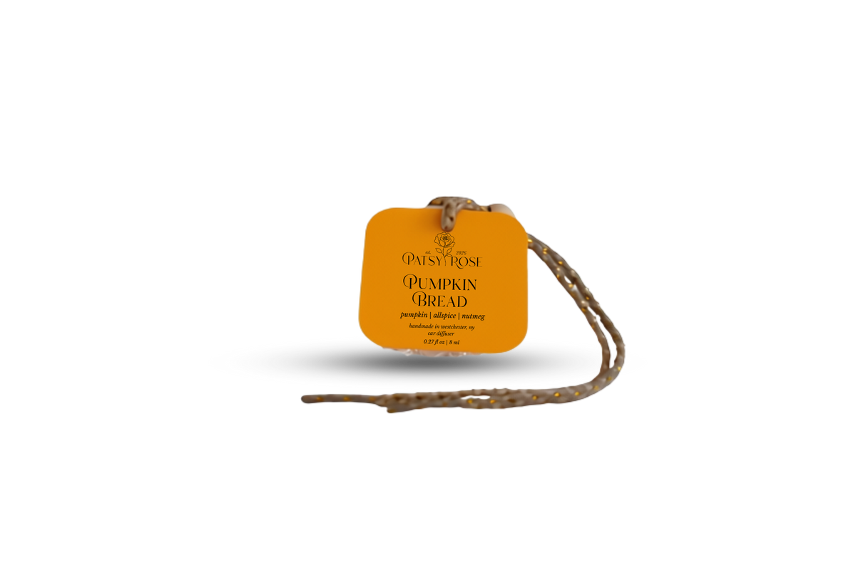An orange-colored paper hang tag with black text reading 'Patsy Rose Pumpkin Bread' on a glass car air freshener, with a braided string attached, isolated on a white background.