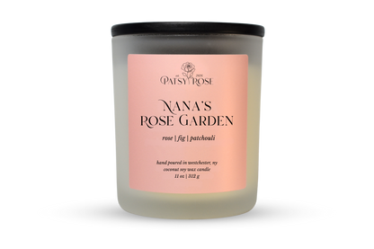 A 11 oz soy wax candle by Patsy Rose in the 'Nana's Rose Garden' 'rose | fig | patchouli' scent. The candle is in a frosted glass jar with a black lid and a peach-pink label, set against a plain white background.