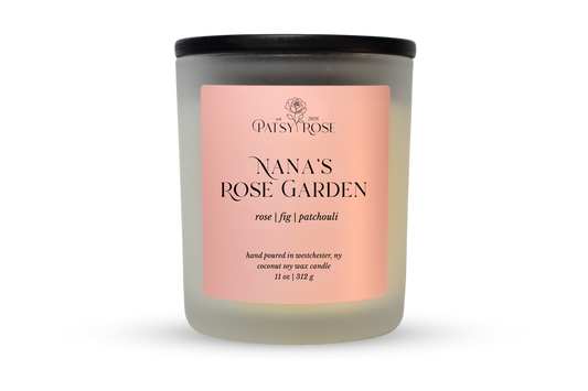 A 11 oz soy wax candle by Patsy Rose in the 'Nana's Rose Garden' 'rose | fig | patchouli' scent. The candle is in a frosted glass jar with a black lid and a peach-pink label, set against a plain white background.