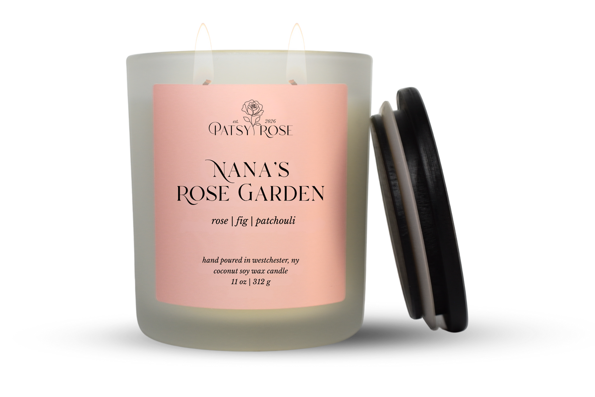An 11 oz double-wick candle in a frosted glass jar with a pale pink label reading 'Patsy Rose Nana's Rose Garden' and 'rose | fig | patchouli.' A black lid is leaning against the right side of the jar on a clean white background.