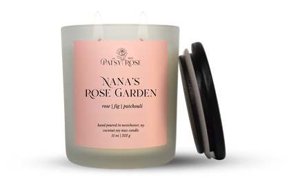 An 11 oz double-wick candle in a frosted glass jar with a pale pink label reading 'Patsy Rose Nana's Rose Garden' and 'rose | fig | patchouli.' A black lid is leaning against the right side of the jar on a clean white background.