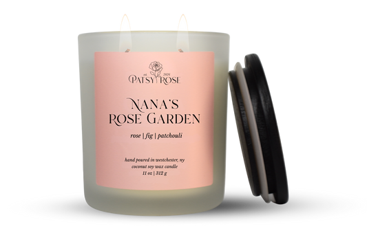 An 11 oz double-wick candle in a frosted glass jar with a pale pink label reading 'Patsy Rose Nana's Rose Garden' and 'rose | fig | patchouli.' A black lid is leaning against the right side of the jar on a clean white background.