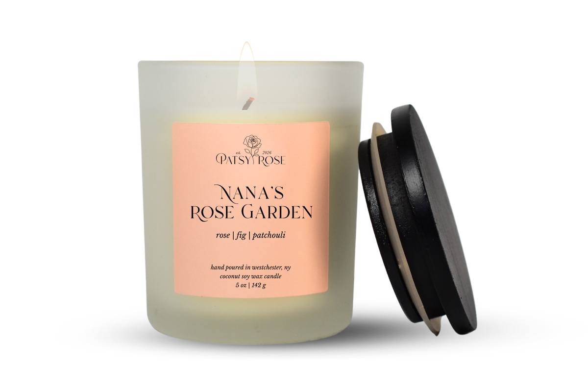 A single-wick coconut soy candle in a frosted glass jar with a pale pink label reading 'Patsy Rose Nana's Rose Garden' and 'rose | fig | patchouli.' A black lid leans against the right side of the jar on a clean white background.