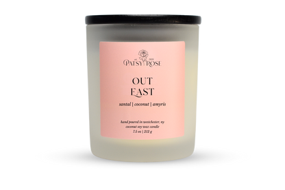 A 7.5 oz soy wax candle by Patsy Rose in the 'Out East' 'santal | coconut | amyris' scent. The candle is in a frosted glass jar with a black lid and a peach-pink label, set against a plain white background.