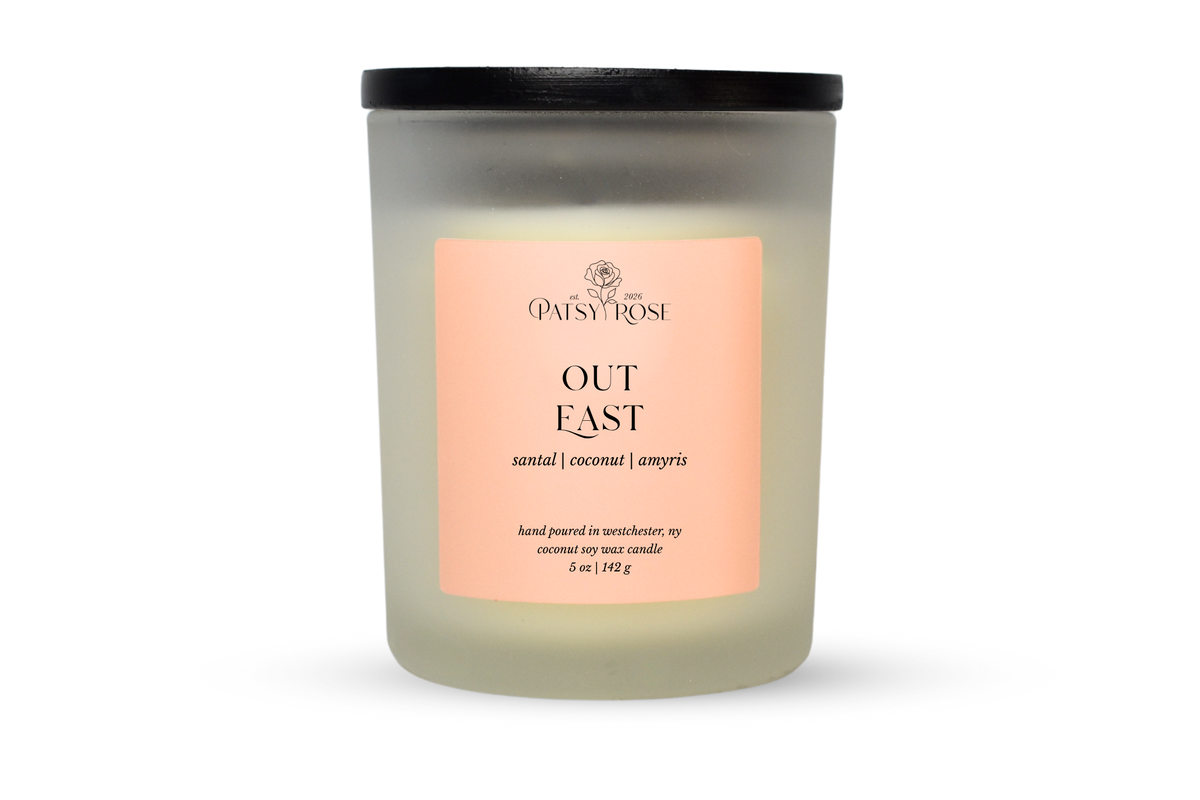 A 5 oz soy wax candle by Patsy Rose in the 'Out East' 'santal | coconut | amyris' scent. The candle is in a frosted glass jar with a black lid and a peach-pink label, set against a plain white background.