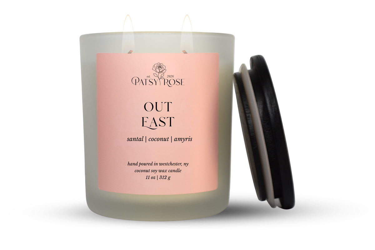 An 11 oz double-wick candle in a frosted glass jar with a pale pink label reading 'Patsy Rose Out East' and 'santal | coconut | amyris.' A black lid is leaning against the right side of the jar on a clean white background.