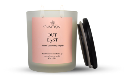 An 11 oz double-wick candle in a frosted glass jar with a pale pink label reading 'Patsy Rose Out East' and 'santal | coconut | amyris.' A black lid is leaning against the right side of the jar on a clean white background.