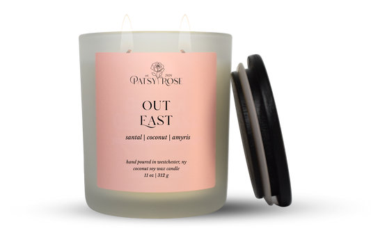 An 11 oz double-wick candle in a frosted glass jar with a pale pink label reading 'Patsy Rose Out East' and 'santal | coconut | amyris.' A black lid is leaning against the right side of the jar on a clean white background.