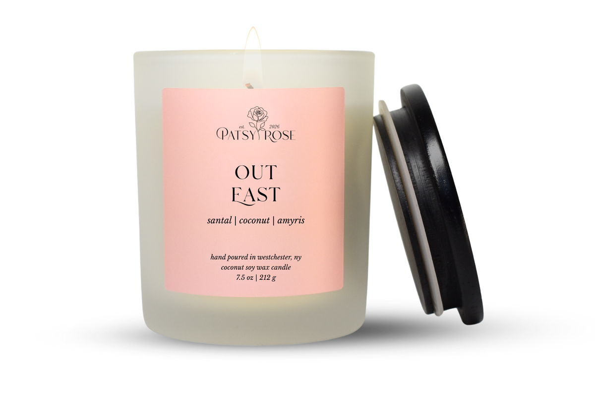 A 7.5 oz single-wick candle in a frosted glass jar with a pale pink label reading 'Patsy Rose Out East' and 'santal | coconut | amyris.' A black lid is leaning against the right side of the jar on a clean white background.