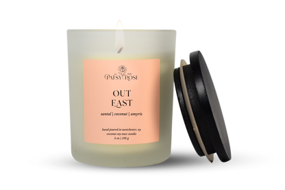 A single-wick coconut soy candle in a frosted glass jar with a pale pink label reading 'Patsy Rose Out East' and 'santal | coconut | amyris.' A black lid leans against the right side of the jar on a clean white background.