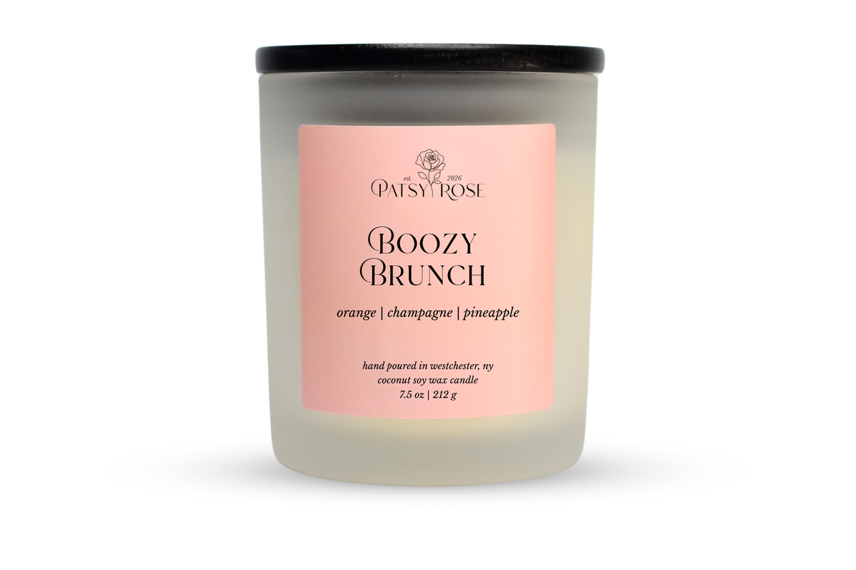 A 7.5 oz soy wax candle by Patsy Rose in the 'Boozy Brunch' 'orange | champagne | pineapple' scent. The candle is in a frosted glass jar with a black lid and a peach-pink label, set against a plain white background.
