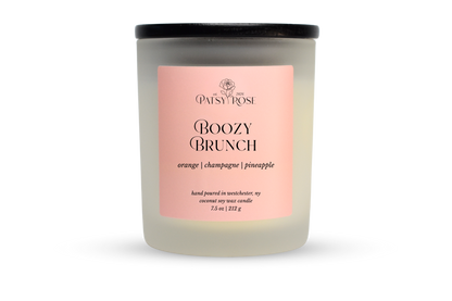 A 7.5 oz soy wax candle by Patsy Rose in the 'Boozy Brunch' 'orange | champagne | pineapple' scent. The candle is in a frosted glass jar with a black lid and a peach-pink label, set against a plain white background.