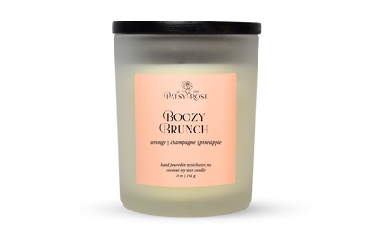 A 5 oz soy wax candle by Patsy Rose in the 'Boozy Brunch' 'orange| champagne | pineapple' scent. The candle is in a frosted glass jar with a black lid and a peach-pink label, set against a plain white background.