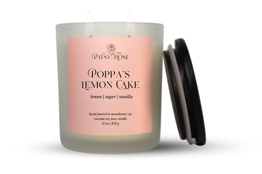 An 11 oz double-wick candle in a frosted glass jar with a pale pink label reading 'Patsy Rose Poppa's Lemon Cake' and 'lemon | sugar | vanilla.' A black lid is leaning against the right side of the jar on a clean white background.