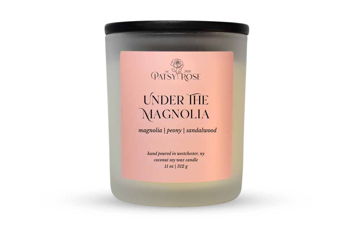 A 11 oz soy wax candle by Patsy Rose in the 'Under The Magnolia' 'magnolia | peony | sandalwood' scent. The candle is in a frosted glass jar with a black lid and a peach-pink label, set against a plain white background.