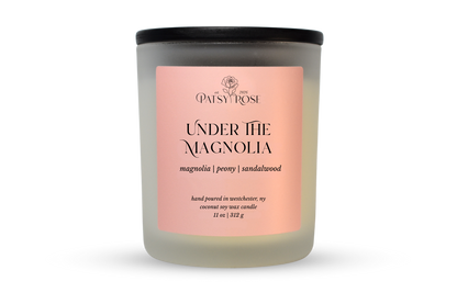 A 11 oz soy wax candle by Patsy Rose in the 'Under The Magnolia' 'magnolia | peony | sandalwood' scent. The candle is in a frosted glass jar with a black lid and a peach-pink label, set against a plain white background.
