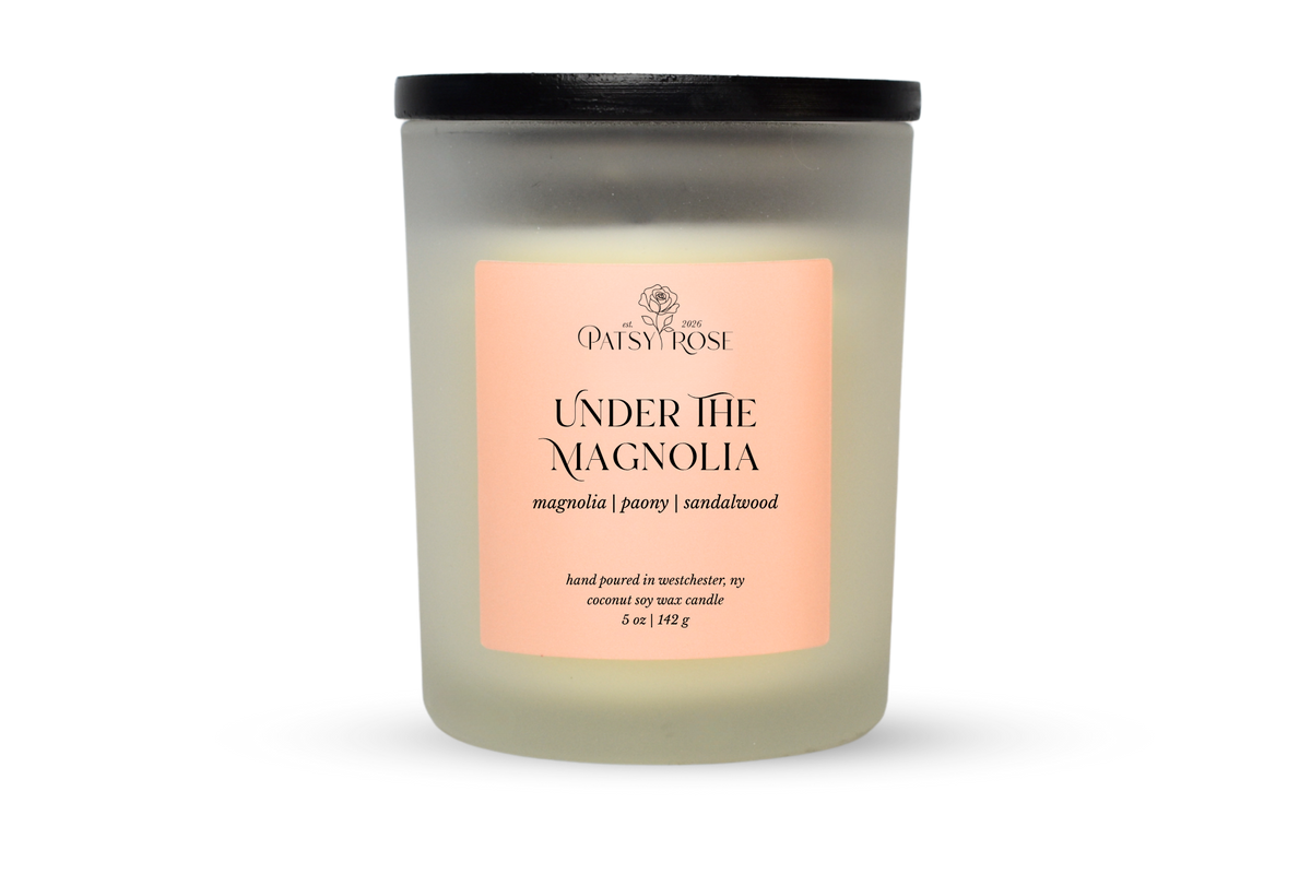 A 5 oz soy wax candle by Patsy Rose in the 'Under The Magnolia' 'magnolia | peony | sandalwood' scent. The candle is in a frosted glass jar with a black lid and a peach-pink label, set against a plain white background.