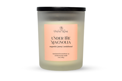 A 5 oz soy wax candle by Patsy Rose in the 'Under The Magnolia' 'magnolia | peony | sandalwood' scent. The candle is in a frosted glass jar with a black lid and a peach-pink label, set against a plain white background.